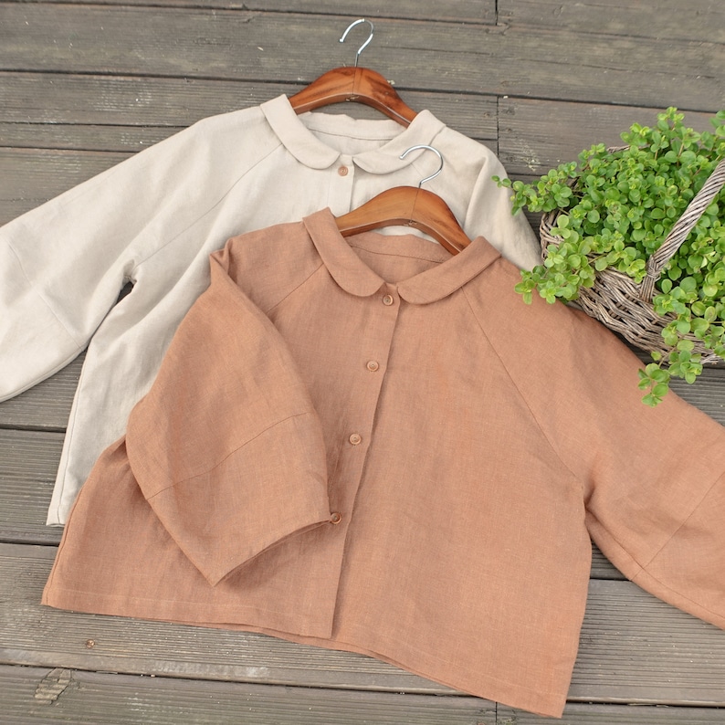 May include: Two linen shirts with peter pan collars, one beige and one brown, hanging on wooden hangers.