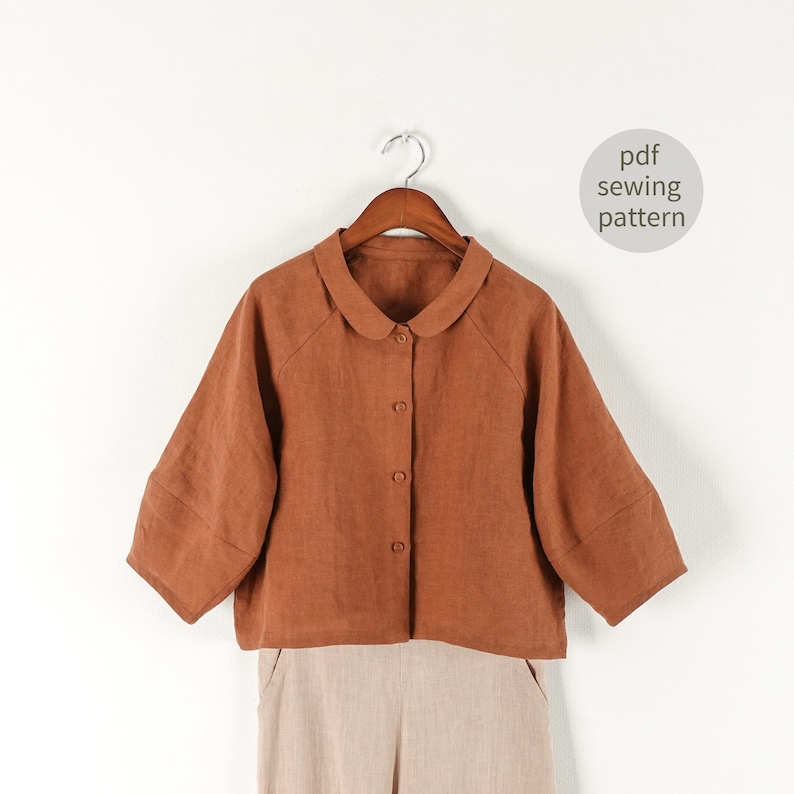May include: A rust-colored linen shirt with a peter pan collar and button closure. The shirt has a cropped length and 3/4 sleeves. The shirt is hanging on a wooden hanger against a white background. The text "pdf sewing pattern" is written in a circle on the right side of the image.