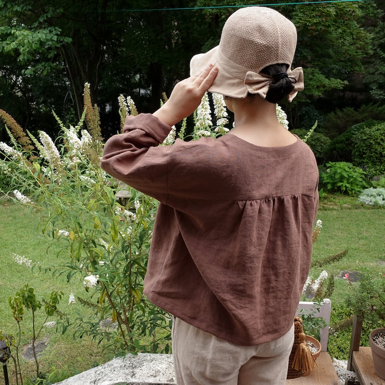 Op de afbeelding: Een bruin linnen blouse met een geplooide achterkant. De blouse heeft lange mouwen en een ontspannen pasvorm. Het model draagt een beige hoed met een strik.