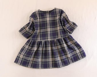 May include: A short-sleeved dress featuring a dark blue, white, and thin yellow plaid pattern. The garment has a round neckline, a button closure at the back of the neck, and a gathered drop-waist skirt that creates a flared, voluminous silhouette.