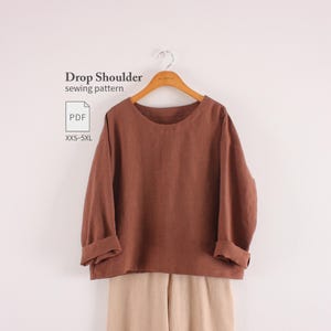 May include: A brown long-sleeve top with a drop shoulder design hanging on a wooden hanger. The text "Drop Shoulder sewing pattern" and "PDF XXS-5XL" is visible on the left. The top is paired with light beige trousers visible at the bottom.