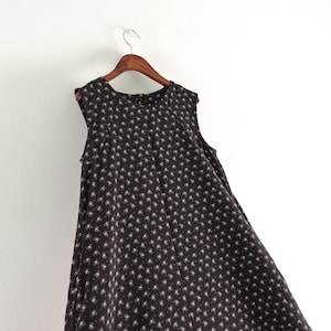 May include: A sleeveless black dress featuring a repeating pattern of small white and pink floral motifs. The garment has a round neckline, a structured bodice with subtle vertical pleats, and a flared skirt silhouette hanging on a wooden hanger.