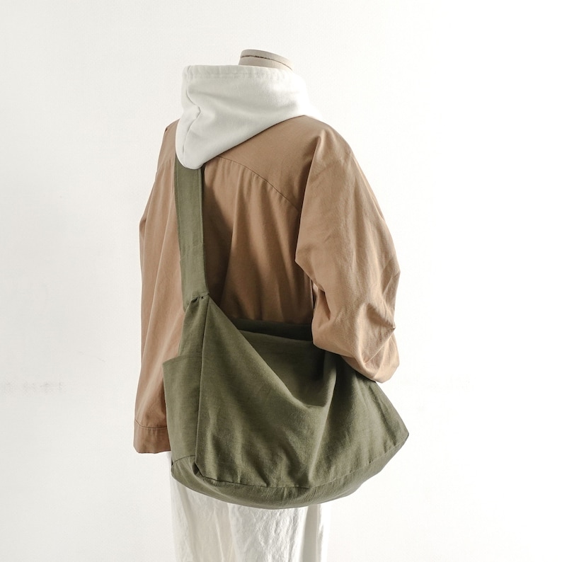May include: A large, olive green canvas tote bag with a long strap. The bag is worn over the shoulder of a mannequin wearing a brown shirt and a white hooded sweatshirt.