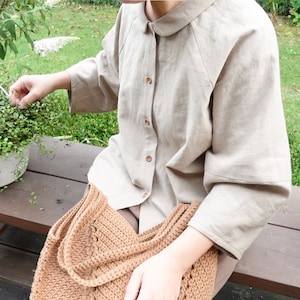 May include: A beige linen shirt with a collared neckline and button closure. The shirt has a relaxed fit and three-quarter sleeves. The person is wearing a brown crocheted bag.