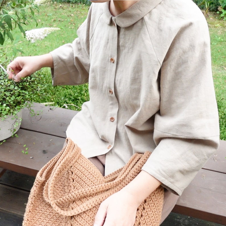 May include: A beige linen shirt with a collared neckline and button closure. The shirt has a relaxed fit and three-quarter sleeves. The person is wearing a brown crocheted bag.