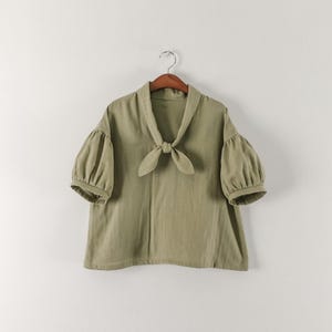 May include: A sage green blouse featuring short puff sleeves with gathered cuffs, a wide sailor-style collar, and a front tie closure at the neckline. The garment is displayed on a wooden hanger against a plain, neutral-coloured background.