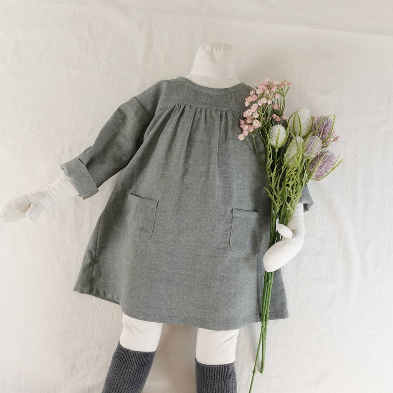 May include: A charcoal grey dress with long sleeves, a round neckline, and two front pockets. The dress features a gathered bodice and is displayed on a mannequin wearing white leggings and dark grey socks, holding a bouquet of pink and green flowers.