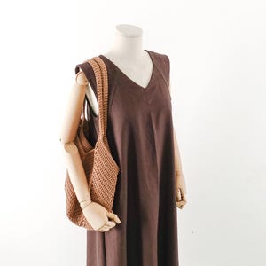 Könnte beinhalten: Ein ärmelloses, dunkelbraunes Kleid mit V-Ausschnitt und eine strukturierte, hellbraune Häkeltasche mit zwei Schulterriemen. Das Kleid weist sichtbare Nahtdetails entlang des Oberteils auf und hat eine gerade, lockere Passform.