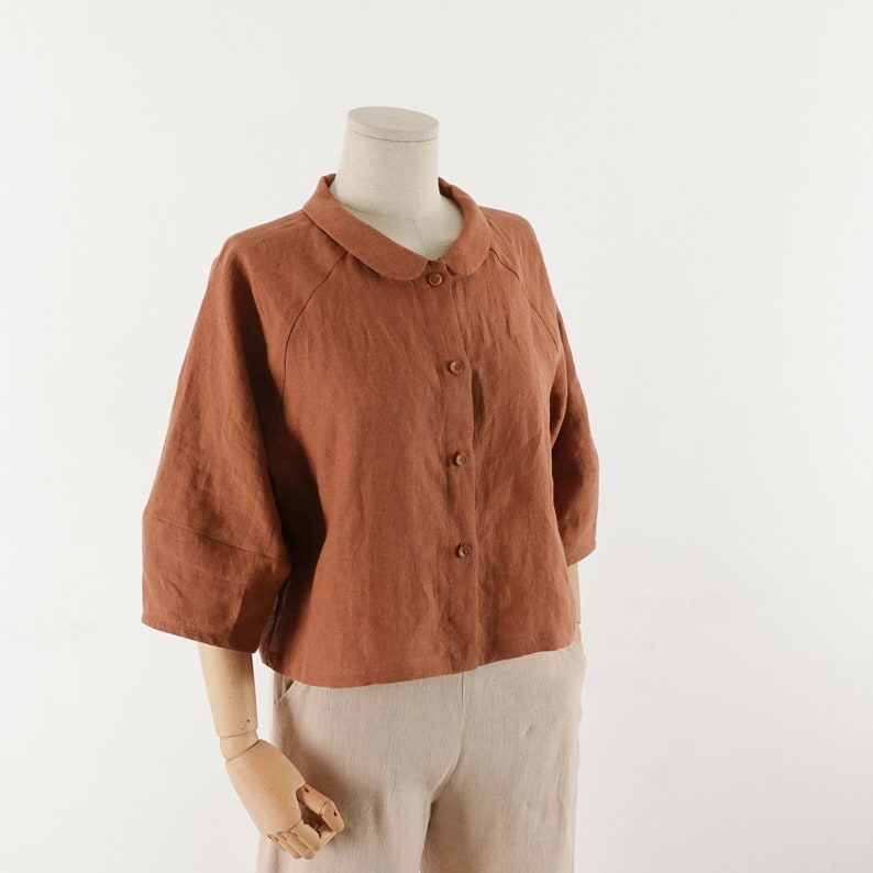 May include: A rust-colored linen shirt with a peter pan collar and button closure. The shirt has a boxy fit and three-quarter sleeves.
