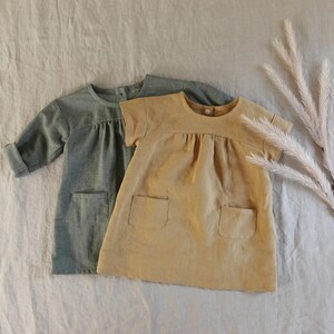 May include: Two short-sleeved dresses, one in sage green and one in mustard yellow. Each dress features a round neckline with a single button closure at the back, a gathered bodice, and two square patch pockets on the front of the skirt.