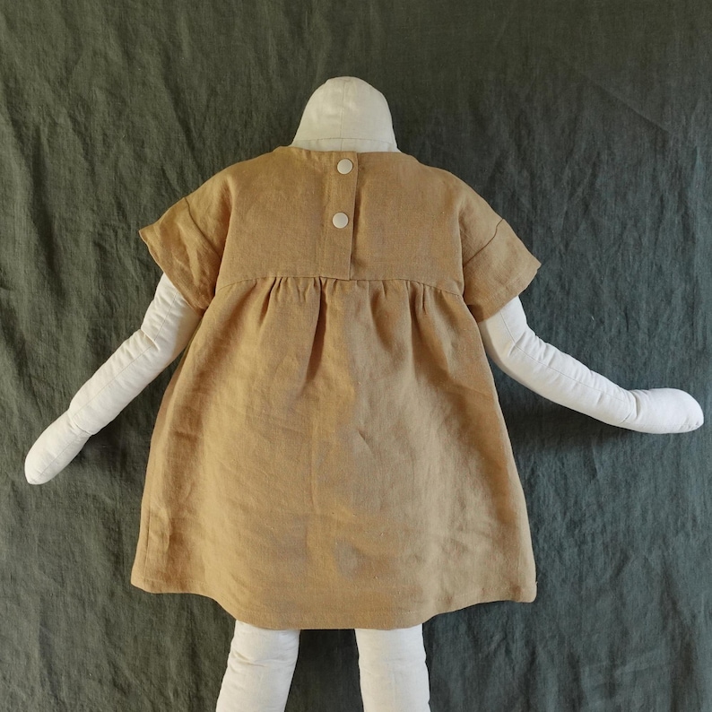May include: A short-sleeved, tan-coloured dress featuring a gathered waistline and a back closure with two white snap buttons. The garment is displayed on a white mannequin form, showcasing a simple, loose-fitting silhouette with a plain texture.
