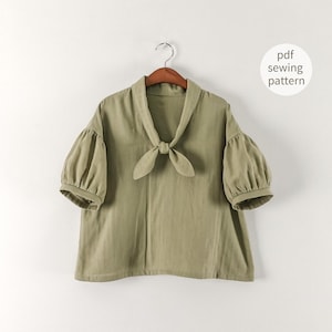 May include: An olive green blouse with short, puffed sleeves and a tie-neck detail. The blouse is hanging on a wooden hanger. Text on the image reads "pdf sewing pattern".