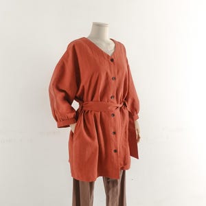 May include: A burnt orange long-sleeve tunic featuring a V-neckline, a front button placket with six dark buttons, and a matching fabric tie belt at the waist. The garment has loose, voluminous sleeves with cuffs and two side pockets.