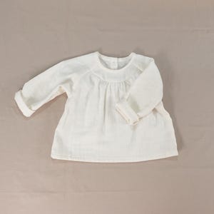 May include: A long-sleeved, off-white shirt featuring a round neckline with subtle gathering at the chest. The garment has cuffed sleeves and a button closure at the back of the neck, constructed from a textured, lightweight fabric with a loose fit.
