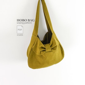 Könnte beinhalten: Eine senfgelbe Hobo-Tasche mit einer Schleife auf der Vorderseite und einem einzelnen Schulterriemen. Der Text links lautet HOBO BAG sewing pattern, PDF und 3 SIZES. Die Tasche hat einen geschwungenen Boden und eine weiche Stoffoptik.