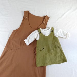Op de afbeelding: Een grote bruine mouwloze jurk met brede banden en twee voorzakken, gecombineerd met een kleinere olijfgroene mouwloze jurk met een wit shirt met lange mouwen eronder. Beide jurken hebben zichtbare knopen op de schouderbanden.