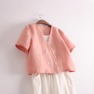 May include: A short-sleeved, double-breasted jacket in a soft peach color with a V-neckline and four white buttons, layered over a plain white garment. The jacket features a structured silhouette with a clean, simple design and short sleeves.