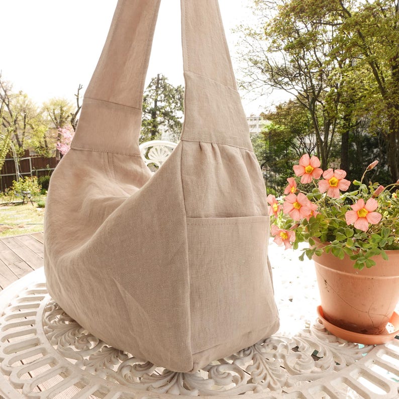 May include: A beige hobo bag crafted from a textured fabric, featuring a wide shoulder strap, a soft slouchy silhouette, and a visible exterior side pocket. The bag has a simple construction with gathered detailing near the top strap attachment.