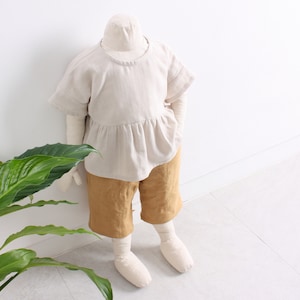 Pode incluir: Uma blusa de linho bege com cintura franzida e mangas curtas, combinada com cal&ccedil;&otilde;es de linho amarelo mostarda. O conjunto &eacute; apresentado num manequim branco.