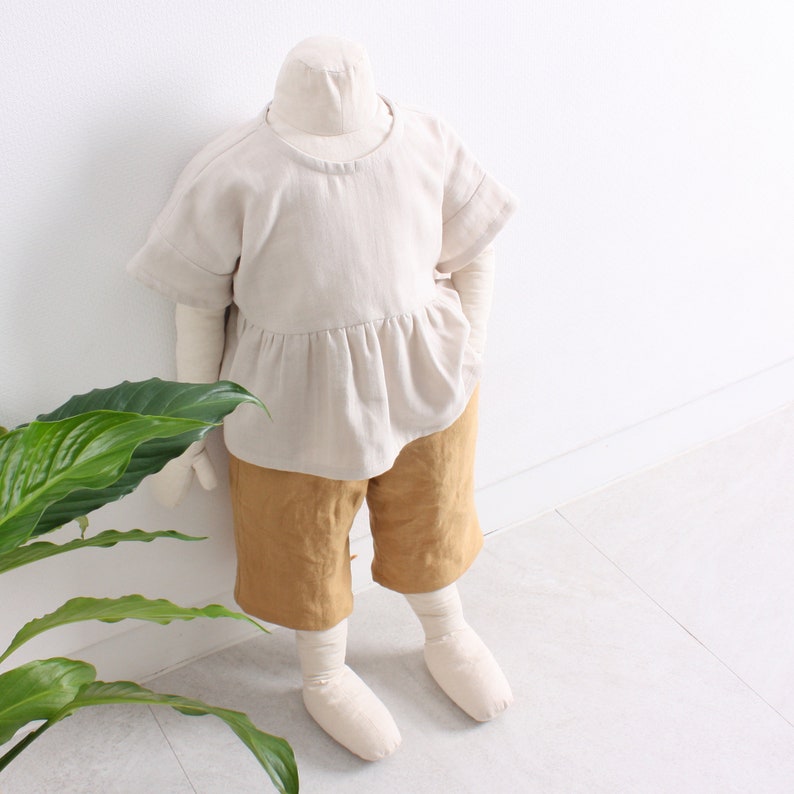 Pode incluir: Uma blusa de linho bege com cintura franzida e mangas curtas, combinada com cal&ccedil;&otilde;es de linho amarelo mostarda. O conjunto &eacute; apresentado num manequim branco.