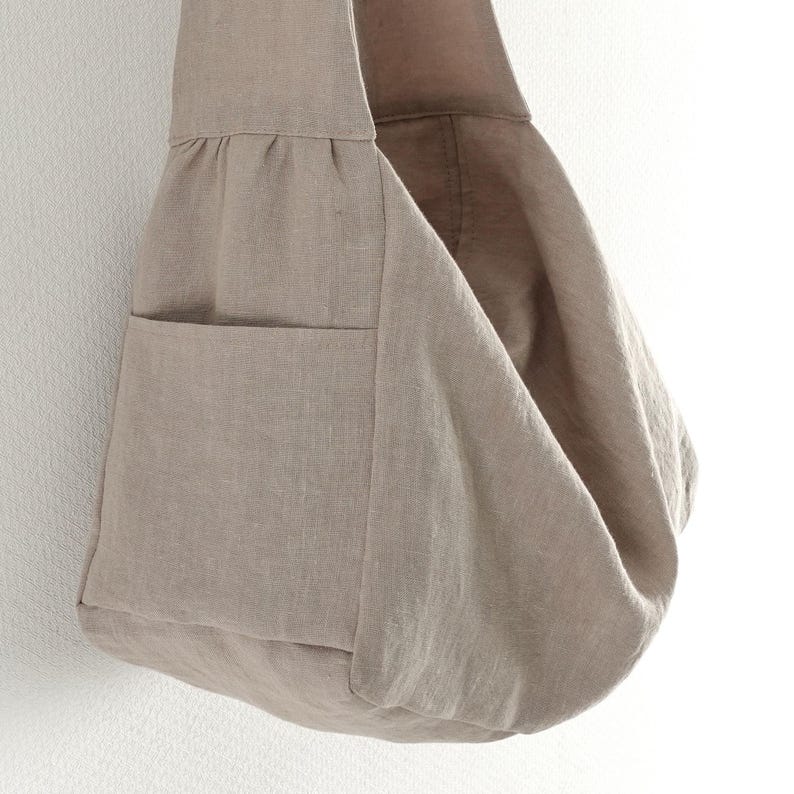 May include: A taupe-colored hobo bag crafted from a textured fabric, featuring a wide shoulder strap, a gathered seam detail along the upper section, and a single exterior pocket on the side, showcasing a soft, unstructured, and slouchy silhouette.
