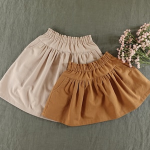 May include: Two gathered skirts, one beige and one brown, with elastic waistbands. The skirts are made of a soft, lightweight fabric.