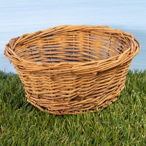 May include: A round, open-weave wicker basket. The basket is made of light brown woven material and sits on a bed of green grass. The basket's sides curve gently outward, creating a shallow bowl shape.
