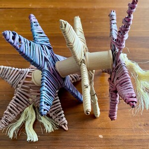 May include: Three woven star-shaped toys in purple, pink, and natural colors with a wooden dowel connecting them. Each star has a long, fluffy tail.
