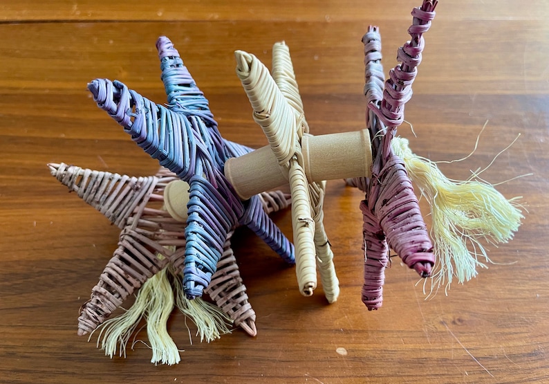 May include: Three woven star-shaped toys in purple, pink, and natural colors with a wooden dowel connecting them. Each star has a long, fluffy tail.