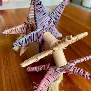 May include: Three woven star-shaped toys with wooden dowels connecting them. The stars are in shades of pink, purple, and beige.