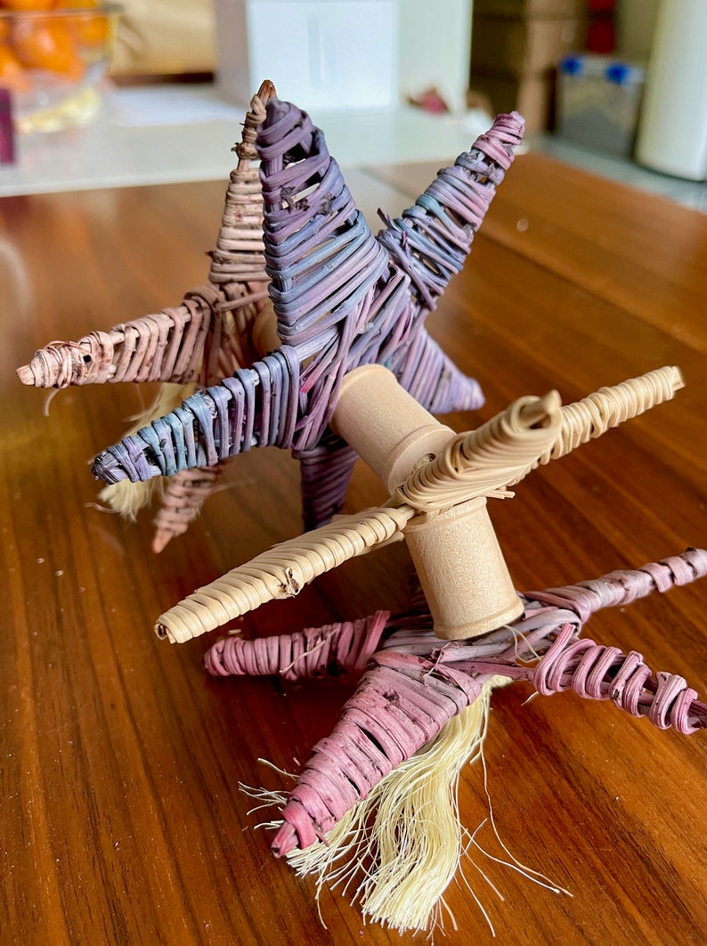 May include: Three woven star-shaped toys with wooden dowels connecting them. The stars are in shades of pink, purple, and beige.