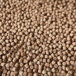 May include: Close-up of a pile of small, brown, pellet-shaped objects. The pellets are evenly distributed and have a slightly textured surface. The overall color is a warm, earthy brown. The image suggests a food product, possibly for pets or livestock.