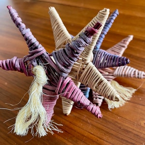 May include: Three woven star ornaments in pink, purple, and beige with a long, white, frayed tassel hanging from the bottom.