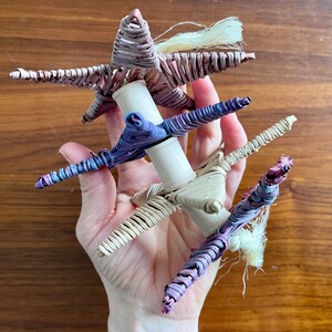 May include: A hand holds a woven star-shaped ornament made of purple, pink, and white straw. The ornament is made of three stars stacked on top of each other.