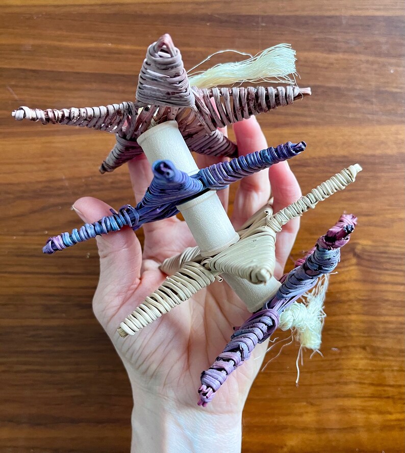 May include: A hand holds a woven star-shaped ornament made of purple, pink, and white straw. The ornament is made of three stars stacked on top of each other.