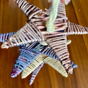 May include: Three woven star ornaments in pink, purple, and natural colors are hanging from a string. The stars are made of thin, woven twigs.