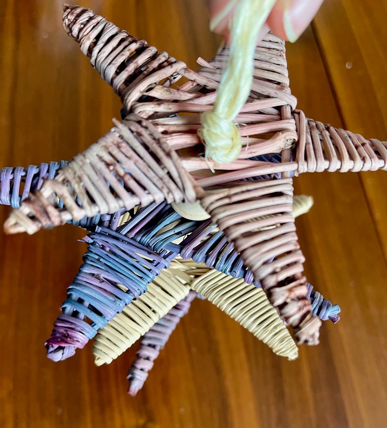 May include: Three woven star ornaments in pink, purple, and natural colors are hanging from a string. The stars are made of thin, woven twigs.