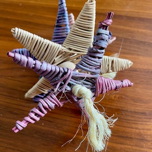 May include: A woven star-shaped ornament with a long, cream-colored tassel. The star is made of natural materials and is painted in shades of purple, pink, and cream.