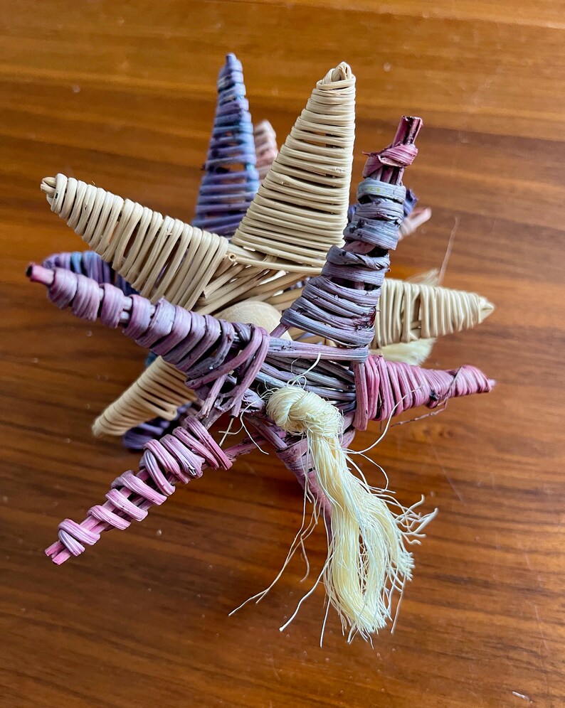 May include: A woven star-shaped ornament with a long, cream-colored tassel. The star is made of natural materials and is painted in shades of purple, pink, and cream.