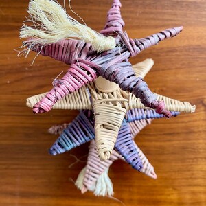 May include: A stack of three woven star ornaments in shades of pink, purple, and beige. The top star has a long, white, frayed tassel.