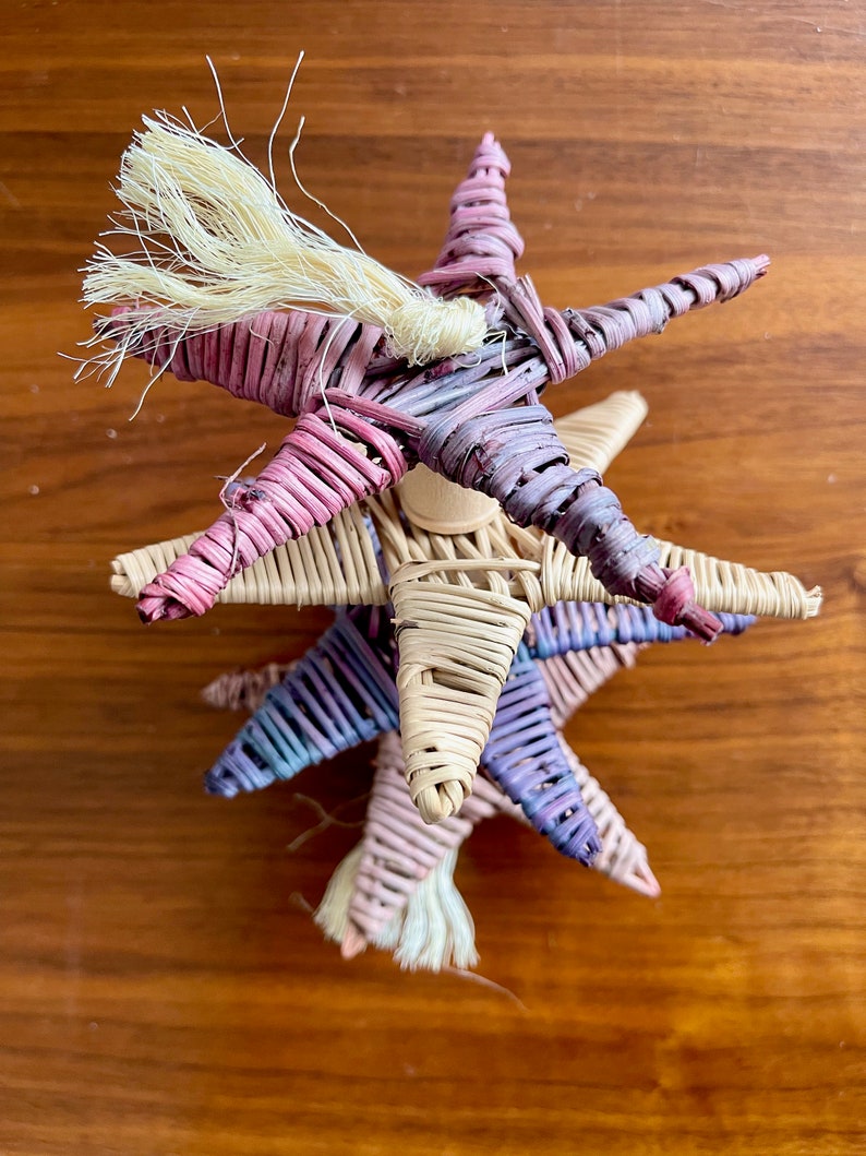 May include: A stack of three woven star ornaments in shades of pink, purple, and beige. The top star has a long, white, frayed tassel.
