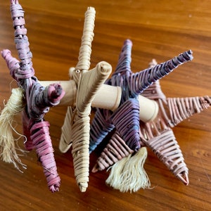 May include: A group of five woven star ornaments with a natural wood base. The stars are made of thin strips of woven material in shades of pink, purple, and beige. Each star has a small, white tassel hanging from the bottom.