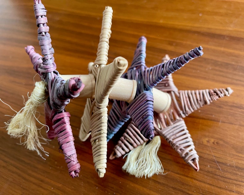 May include: A group of five woven star ornaments with a natural wood base. The stars are made of thin strips of woven material in shades of pink, purple, and beige. Each star has a small, white tassel hanging from the bottom.