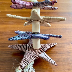 May include: A woven star-shaped ornament with a white tassel. The ornament is made of natural materials and has a rustic look. It is stacked on top of two other similar ornaments, one in purple and one in blue.
