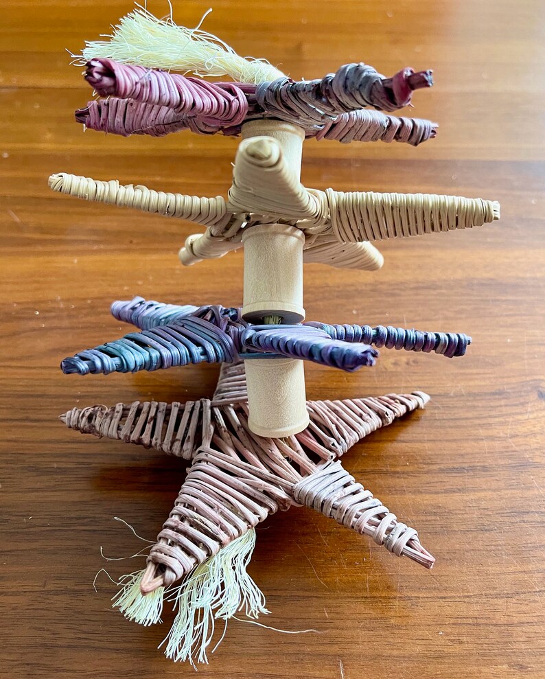 May include: A woven star-shaped ornament with a white tassel. The ornament is made of natural materials and has a rustic look. It is stacked on top of two other similar ornaments, one in purple and one in blue.