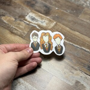 May include: A sticker featuring cartoon illustrations of three characters from the Harry Potter series. The characters are Harry Potter, Ron Weasley, and Hermione Granger. The sticker is white with a black outline and features the characters in their Hogwarts school uniforms.