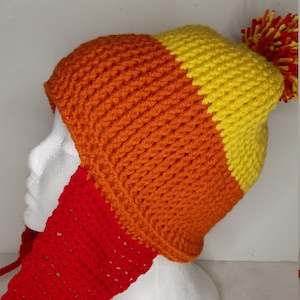 Puede incluir: Un gorro de punto con un esquema de colores rojo, naranja y amarillo. El gorro tiene un pompón en la parte superior y una larga bufanda roja unida a la parte posterior.