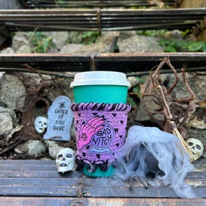 Könnte beinhalten: Ein türkisfarbener Reisebecher mit weißem Deckel und einer lila Häkelmanschette. Die Manschette zeigt ein rosa Handdesign, eine Kristallkugel und die Worte "BAD WITCH". Dekorative Totenköpfe und ein Grabstein mit dem Text "ENTER IF YOU DARE" befinden sich im Hintergrund.
