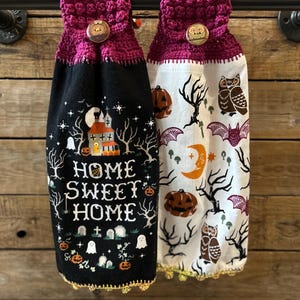 Può includere: Due strofinacci da cucina appesi con parti superiori in uncinetto magenta e bottoni in legno. Uno è nero con una casa stregata, zucche, fantasmi e il testo "HOME SWEET HOME". L'altro è bianco con un motivo di gufi, pipistrelli, zucche e lune.