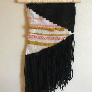 Op de afbeelding: Een geweven wandkleed met een geometrisch ontwerp. Het bovenste gedeelte is zwart, met een diagonale witte en roze sectie. Daaronder bevinden zich horizontale strepen in goud en roze, en een zwarte franje. Het wandkleed hangt aan een houten deuvel.